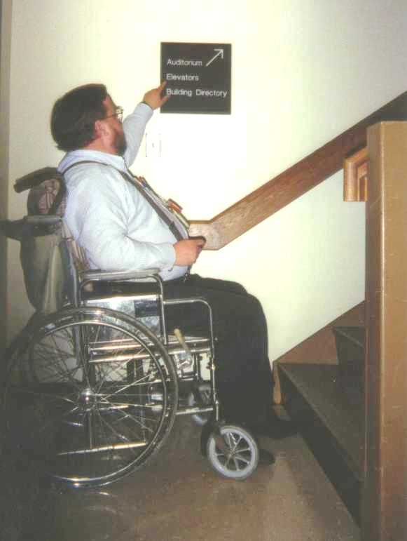 John, in wheelchair, pointing at sign indicating elevators are up a flight of stairs.